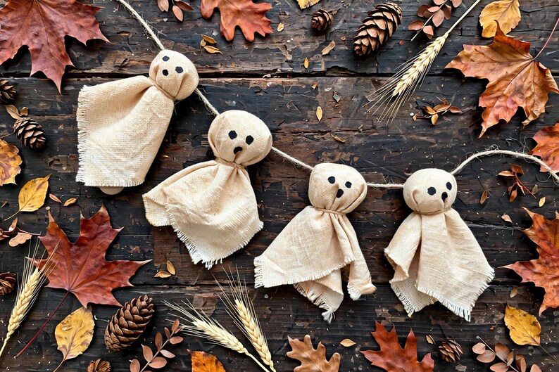May include: A garland featuring four ghost-shaped figures made from light beige fabric with frayed edges. Each ghost has two black circular eyes and a small black circular mouth, all suspended from a single piece of natural-colored twine.