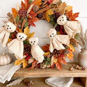 May include: A decorative autumn wreath featuring orange, yellow, and brown maple leaves, pinecones, cinnamon sticks, and red berries. A garland of four small, beige fabric ghost figures with black dot eyes and mouths is draped across the front.