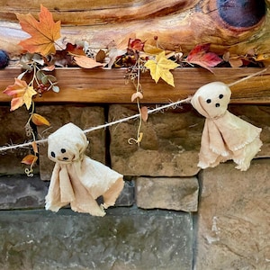May include: A decorative garland featuring two beige fabric ghost figures with black stitched eyes and mouths, suspended on a thin twine string. The ghosts are interspersed with artificial autumn leaves in shades of orange, yellow, and brown.