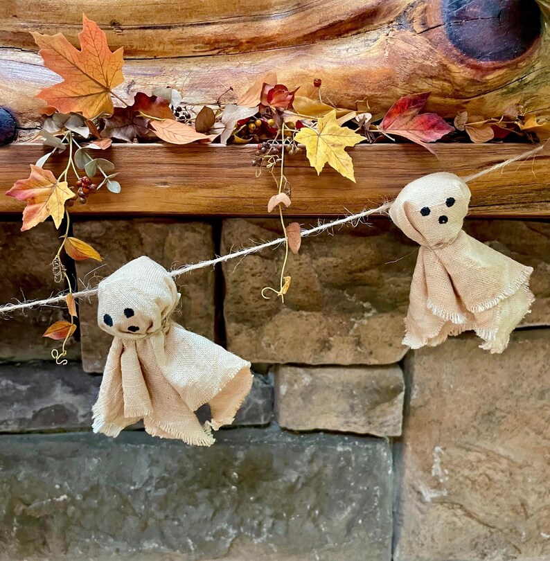 May include: A decorative garland featuring two beige fabric ghost figures with black stitched eyes and mouths, suspended on a thin twine string. The ghosts are interspersed with artificial autumn leaves in shades of orange, yellow, and brown.