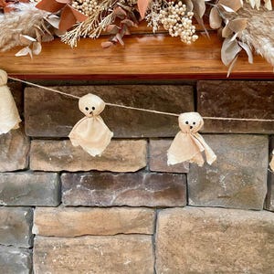 May include: A garland featuring four small ghost figures made from light beige fabric. Each ghost has a rounded head and a draped body with two small black dots for eyes and a single black dot for a mouth, all strung together on a thin twine.