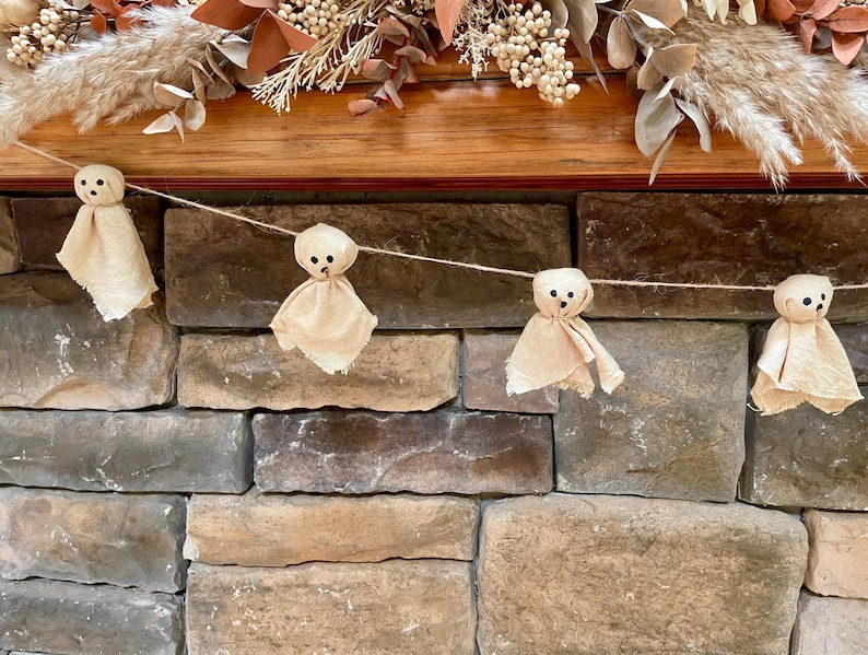 May include: A garland featuring four small ghost figures made from light beige fabric. Each ghost has a rounded head and a draped body with two small black dots for eyes and a single black dot for a mouth, all strung together on a thin twine.