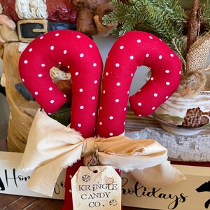 May include: Two red fabric candy canes with white polka dots, tied together with a cream-colored bow. A tag reads "Kringle Candy Co." with a candy cane illustration. Festive holiday decor.