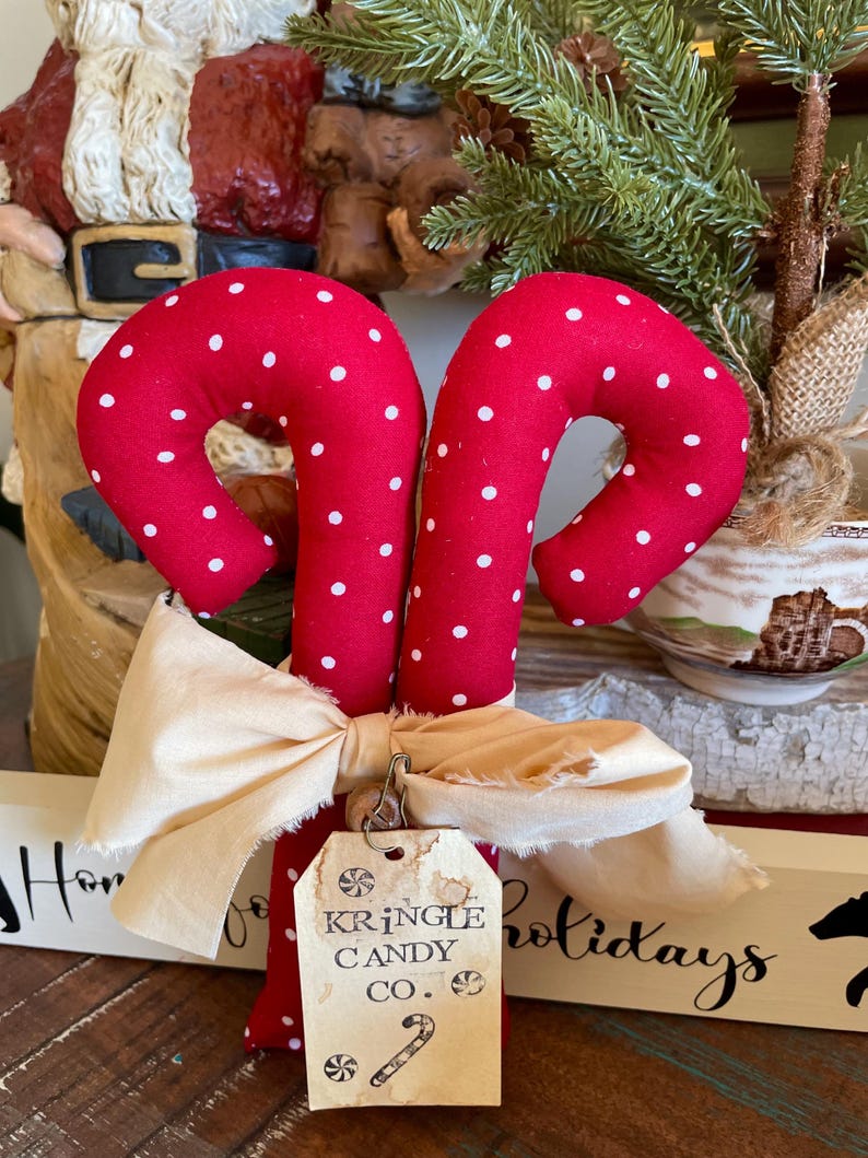 May include: Two red fabric candy canes with white polka dots, tied together with a cream-colored bow. A tag reads "Kringle Candy Co." with a candy cane illustration. Festive holiday decor.