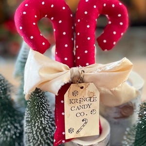 May include: Two red fabric candy canes with white polka dots, tied with a cream-colored ribbon. A tag reads "Kringle Candy Co." with a candy cane illustration. The candy canes are part of a holiday-themed display.
