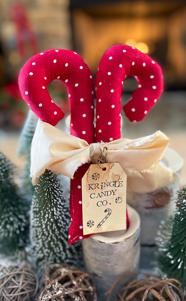 May include: Two red fabric candy canes with white polka dots, tied with a cream-colored ribbon. A tag reads "Kringle Candy Co." with a candy cane illustration. The candy canes are part of a holiday-themed display.