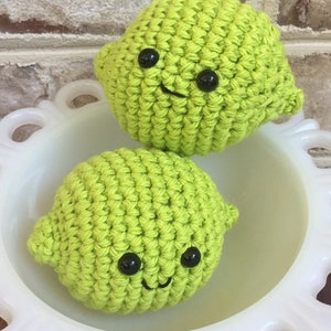 May include: Two crocheted lime-shaped plushies with black button eyes and smiling mouths. The plushies are sitting in a white bowl.