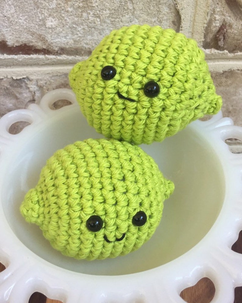 May include: Two crocheted lime-shaped plushies with black button eyes and smiling mouths. The plushies are sitting in a white bowl.