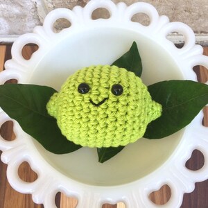 May include: A crocheted lime with a smiling face and black eyes, sitting on a white pedestal with green leaves.