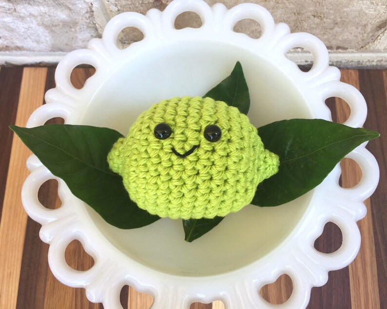 May include: A crocheted lime with a smiling face and black eyes, sitting on a white pedestal with green leaves.