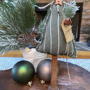 May include: A small, green and white striped fabric Christmas tree with a wooden base. The tree is decorated with a wooden star, a bell, and a tag that says "JOY".