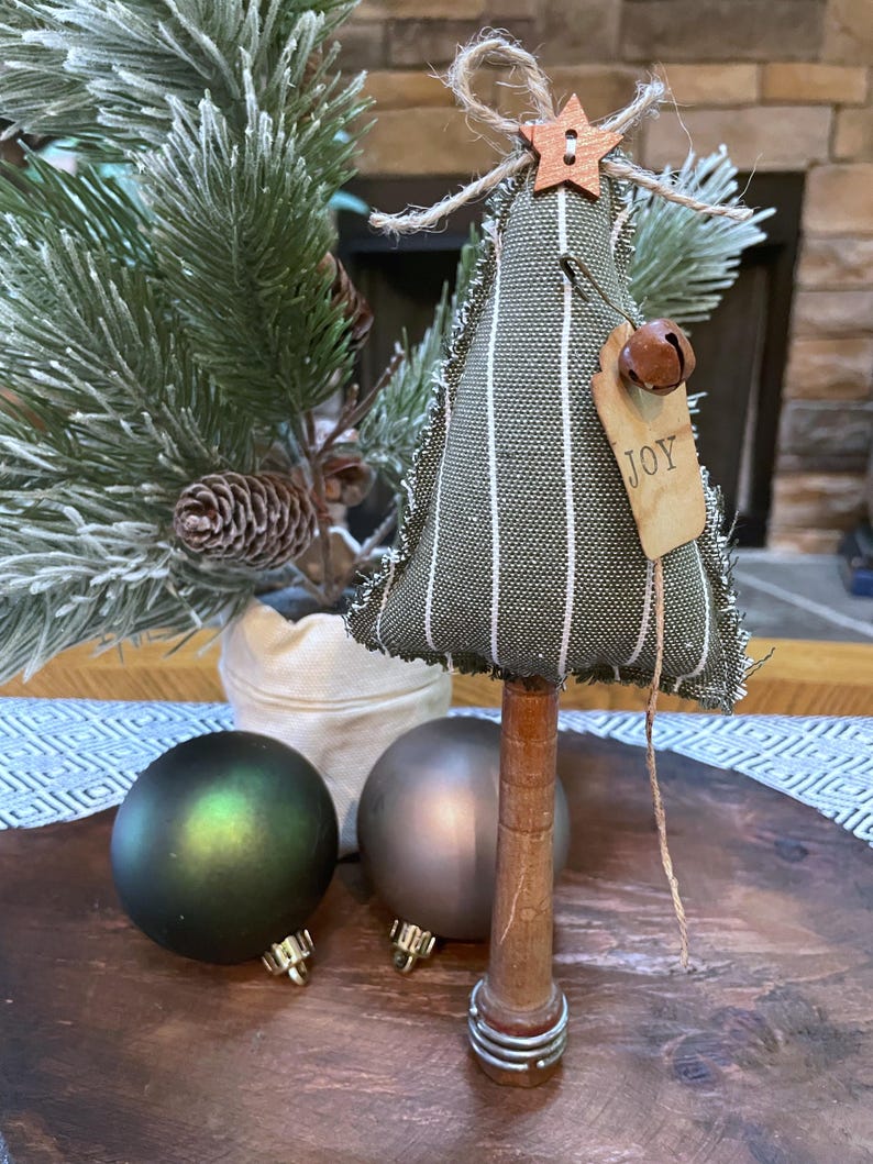 May include: A small, green and white striped fabric Christmas tree with a wooden base. The tree is decorated with a wooden star, a bell, and a tag that says "JOY".