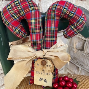 May include: Two fabric candy canes featuring a red, green, and yellow plaid pattern. A beige frayed ribbon is tied around the center, holding a rectangular paper tag with the text MERRY & BRIGHT! and a small metal bell, plus red berries.