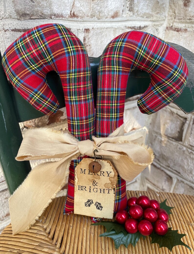 May include: Two fabric candy canes featuring a red, green, and yellow plaid pattern. A beige frayed ribbon is tied around the center, holding a rectangular paper tag with the text MERRY & BRIGHT! and a small metal bell, plus red berries.