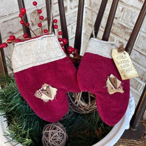 May include: Two red and white fabric mitten ornaments with a heart-shaped button and a tag that reads "Cold Hands Warm Heart".