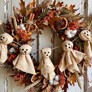May include: A fall wreath featuring dried oak leaves, cinnamon sticks, pinecones, wheat stalks, orange slices, cotton bolls, and red berries. A garland of four beige fabric ghost figures with black stitched eyes and mouths hangs across the center.