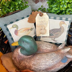 May include: A dark brown wicker basket containing a carved wooden mallard duck decoy, two sage green fabric pillows with printed duck illustrations, and a beige paper tag with the text INTO THE WOODS I GO and a small metal bell attached.