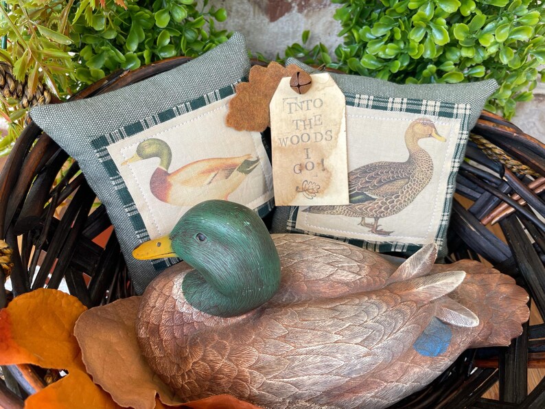May include: A dark brown wicker basket containing a carved wooden mallard duck decoy, two sage green fabric pillows with printed duck illustrations, and a beige paper tag with the text INTO THE WOODS I GO and a small metal bell attached.