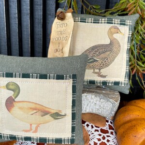 May include: Two square sage green fabric pillows featuring a central cream-colored patch with a printed mallard duck. One pillow includes a tan paper tag with the text INTO THE WOODS I GO and a small metal bell attached to the top corner.