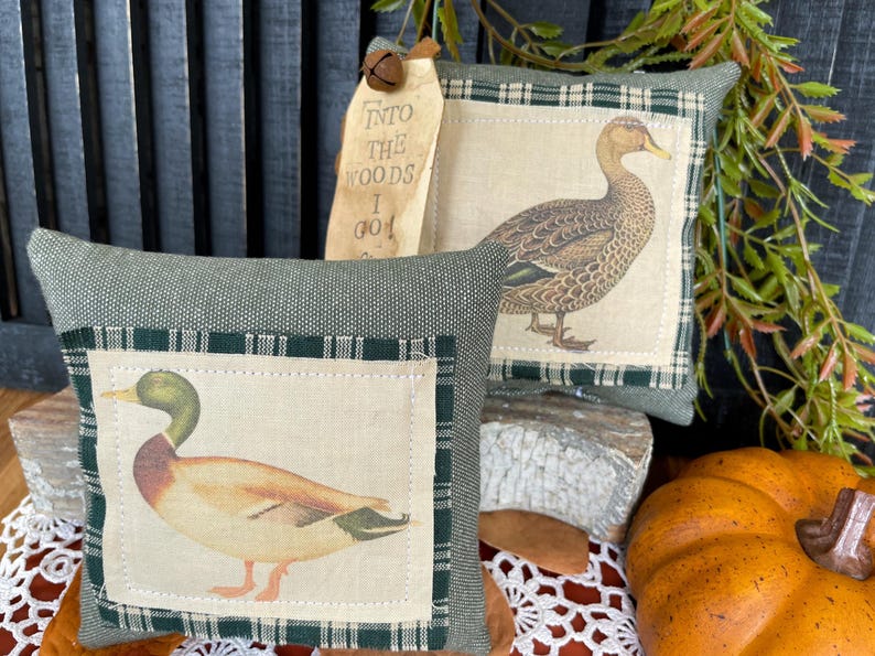 May include: Two square sage green fabric pillows featuring a central cream-colored patch with a printed mallard duck. One pillow includes a tan paper tag with the text INTO THE WOODS I GO and a small metal bell attached to the top corner.