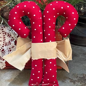 May include: Two red fabric candy canes with white polka dots are tied together with a beige ribbon. The candy canes are a festive holiday decoration, perfect for Christmas. The background includes greenery and a red wooden box.