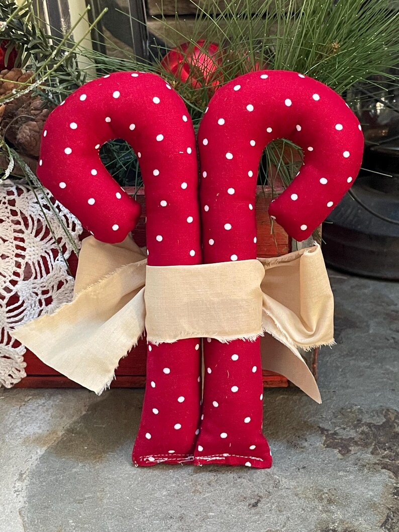 May include: Two red fabric candy canes with white polka dots are tied together with a beige ribbon. The candy canes are a festive holiday decoration, perfect for Christmas. The background includes greenery and a red wooden box.