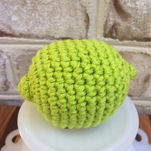 May include: A crocheted lime, green in color, is sitting on a white pedestal. The lime is made with a textured stitch.