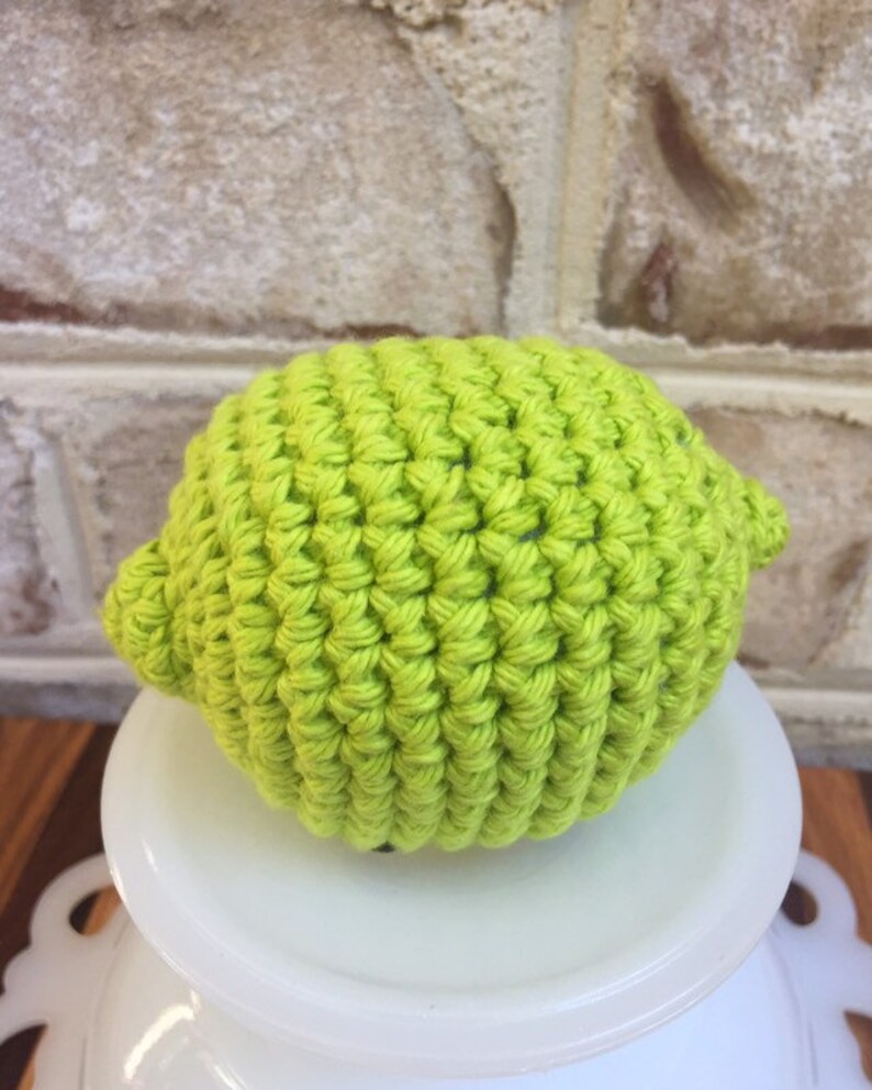 May include: A crocheted lime, green in color, is sitting on a white pedestal. The lime is made with a textured stitch.