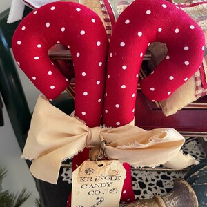 May include: Two red fabric candy canes with white polka dots, tied together with a cream-colored bow. A tag reads "Kringle Candy Co." and features a candy cane illustration. Festive holiday decor.