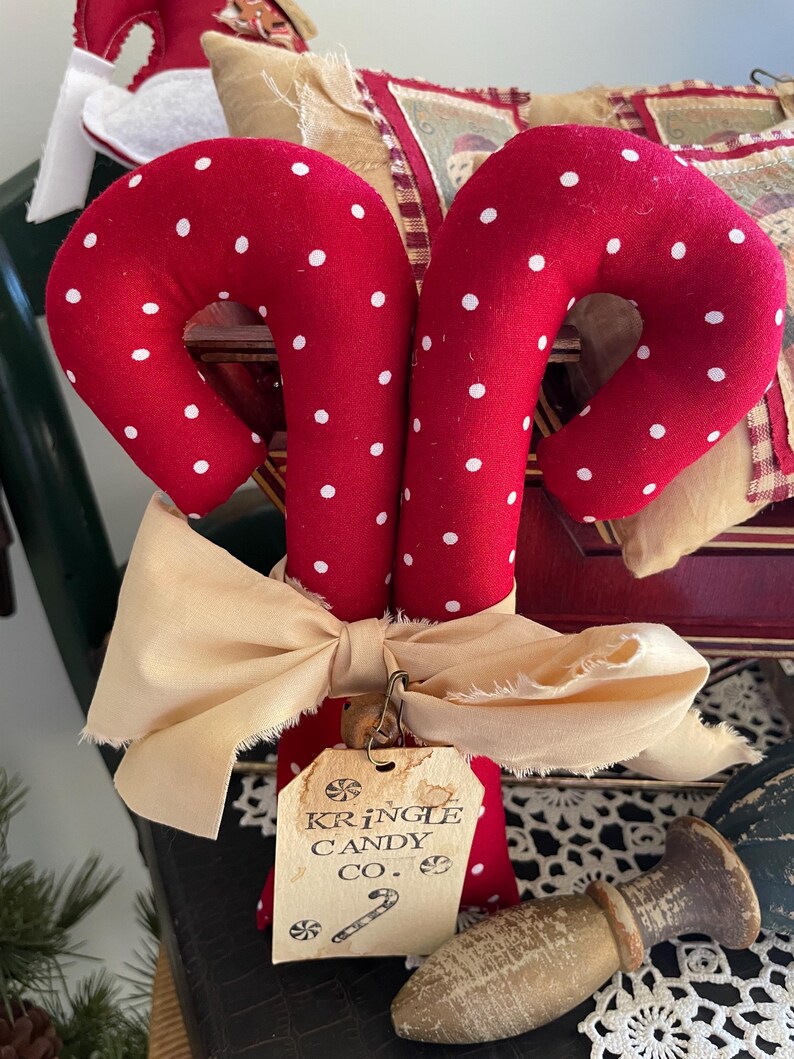 May include: Two red fabric candy canes with white polka dots, tied together with a cream-colored bow. A tag reads "Kringle Candy Co." and features a candy cane illustration. Festive holiday decor.