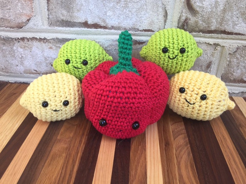 May include: Five crocheted stuffed toys shaped like fruits and vegetables. There are two yellow lemons, one green lime, and one red pepper. All of the toys have black eyes and a smiling mouth.