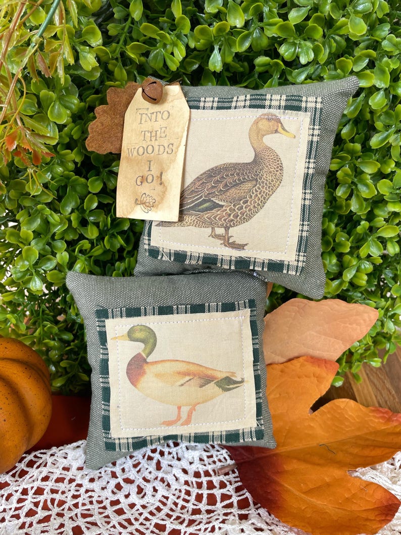May include: Two square olive green fabric pillows featuring centered cream-colored patches with detailed illustrations of ducks. A tan paper tag attached with a small metal bell displays the text INTO THE WOODS I GO in black ink.