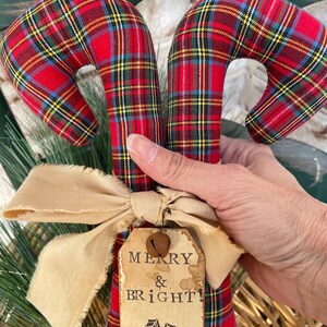 May include: Two fabric candy cane ornaments featuring a red, green, and yellow plaid pattern. A beige frayed ribbon bow is tied around the base, accented by a small metal bell and a rectangular paper tag printed with the text MERRY & BRIGHT.
