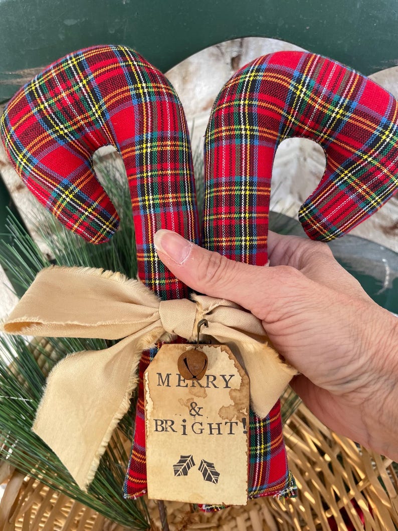 May include: Two fabric candy cane ornaments featuring a red, green, and yellow plaid pattern. A beige frayed ribbon bow is tied around the base, accented by a small metal bell and a rectangular paper tag printed with the text MERRY & BRIGHT.