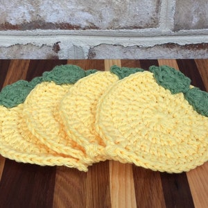 May include: Four crocheted lemon-shaped coasters in yellow with green leaves. The coasters are stacked on top of each other.