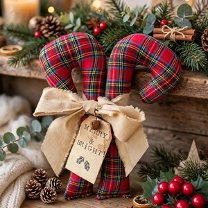 May include: Two fabric candy cane ornaments featuring a red, green, and yellow plaid pattern. The canes are tied together with a wide, frayed beige ribbon and a rectangular paper tag that reads MERRY & BRIGHT! with two small leaf icons.