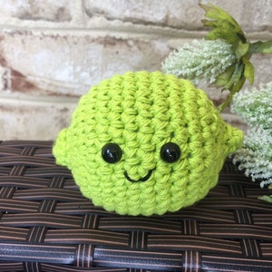 May include: A green crocheted lime with a smiling face and black eyes. The lime is sitting on a brown wicker surface.