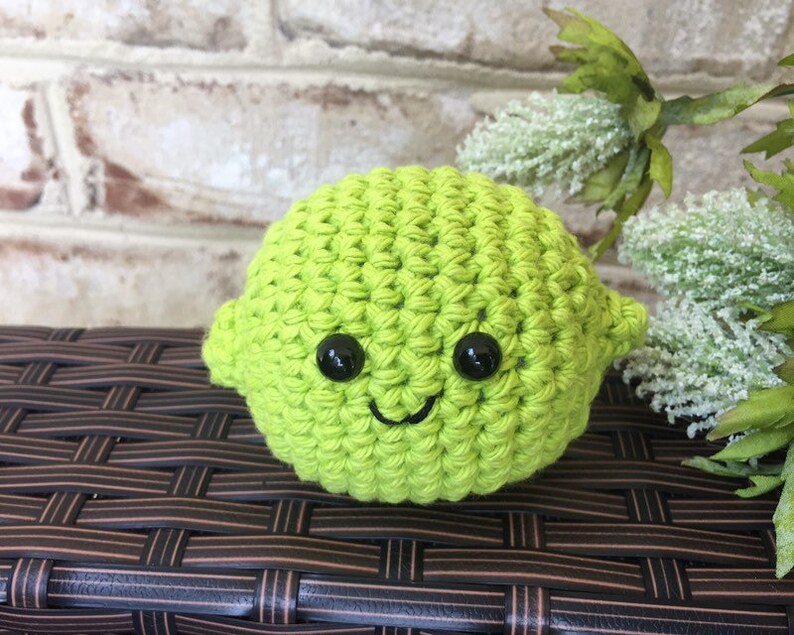 May include: A green crocheted lime with a smiling face and black eyes. The lime is sitting on a brown wicker surface.