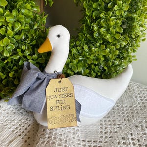 May include: A white fabric goose with a yellow beak and a gray bow tie. A brown tag reads "Just Quackers for Spring" with floral accents. The goose is set against a green wreath.