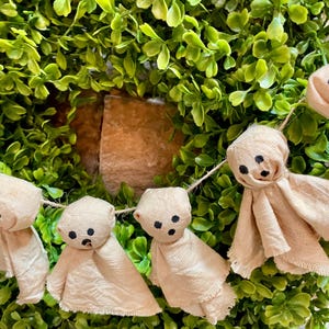 May include: A garland featuring multiple small, beige fabric ghost figures strung together. Each ghost is crafted from light-colored cloth with frayed edges and features three simple black dots representing eyes and a mouth, suspended on a thin twine.