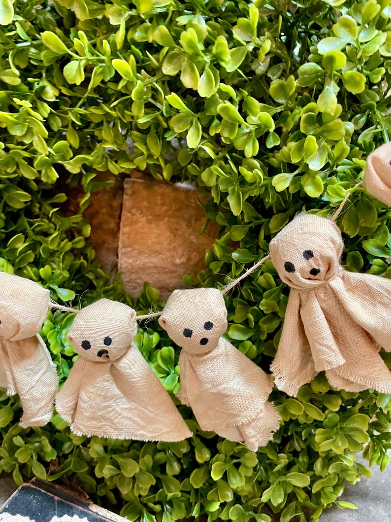 May include: A garland featuring multiple small, beige fabric ghost figures strung together. Each ghost is crafted from light-colored cloth with frayed edges and features three simple black dots representing eyes and a mouth, suspended on a thin twine.