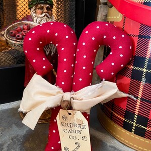 May include: Two red fabric candy canes with white polka dots, tied together with a cream-colored bow. A tag reads "Kringle Candy Co." with a candy cane graphic. A Santa Claus figurine is in the background.