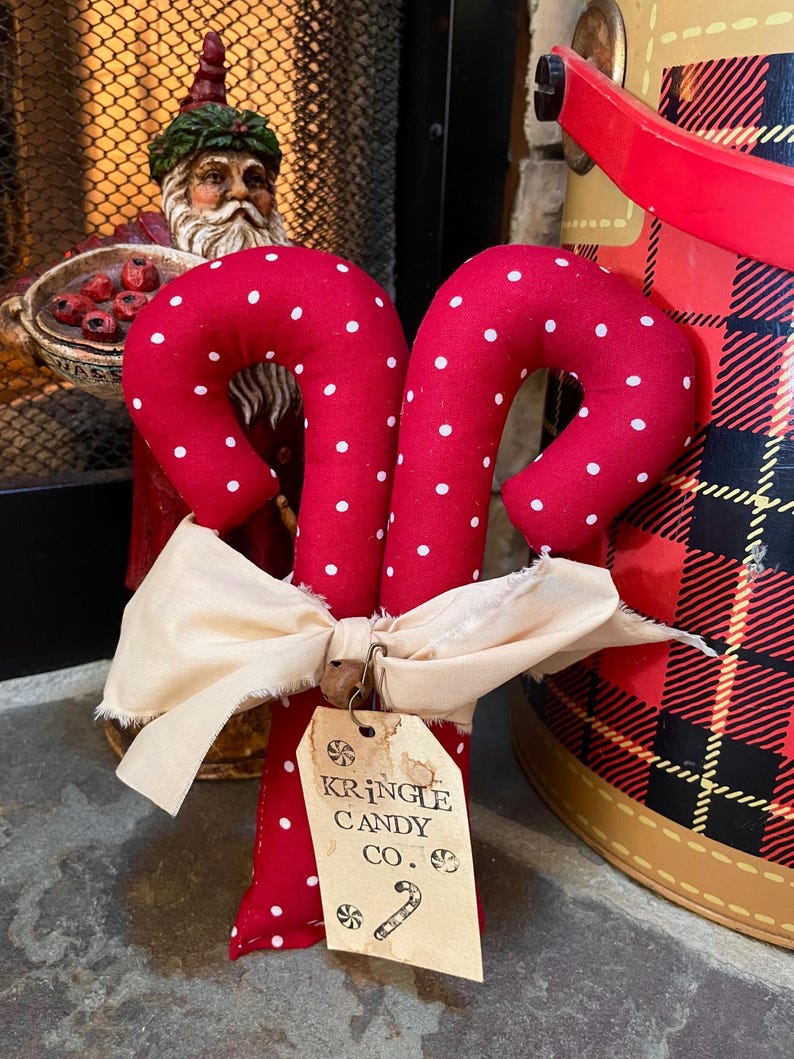 May include: Two red fabric candy canes with white polka dots, tied together with a cream-colored bow. A tag reads "Kringle Candy Co." with a candy cane graphic. A Santa Claus figurine is in the background.