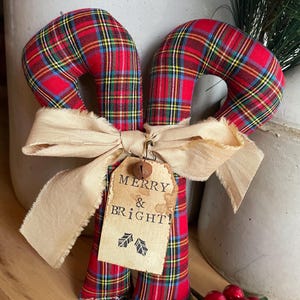 May include: Two fabric candy canes featuring a red, yellow, and blue plaid pattern. A beige frayed ribbon is tied around the center, holding a rectangular paper tag with the text MERRY & BRIGHT! and two holly leaf icons, topped with a small metal bell.