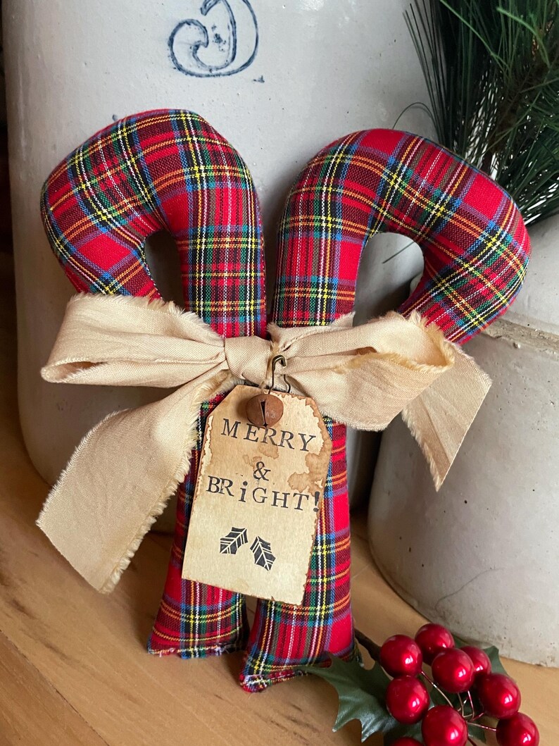 May include: Two fabric candy canes featuring a red, yellow, and blue plaid pattern. A beige frayed ribbon is tied around the center, holding a rectangular paper tag with the text MERRY & BRIGHT! and two holly leaf icons, topped with a small metal bell.