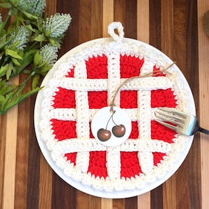 May include: A crocheted cherry pie hot pad with a white border and red center. The hot pad has a loop for hanging. A small tag with two cherries is attached to the hot pad.