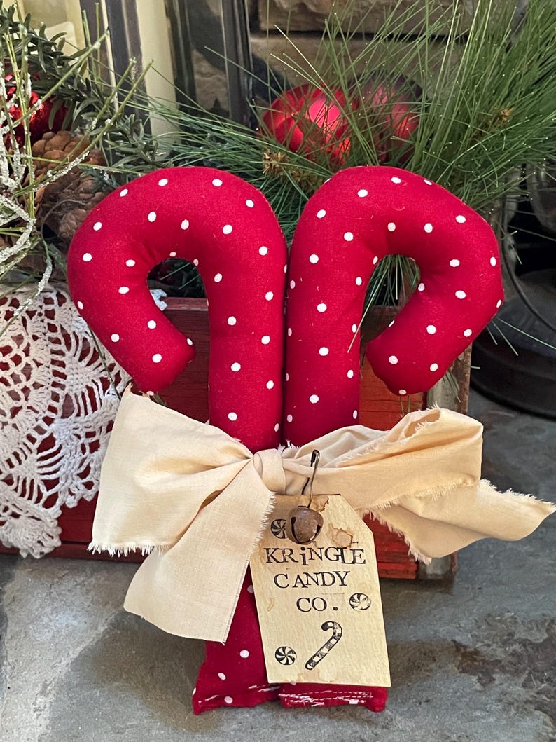 May include: Two red fabric candy canes with white polka dots, tied together with a cream-colored bow. A tag reads "Kringle Candy Co." with a candy cane illustration. Festive holiday decor.