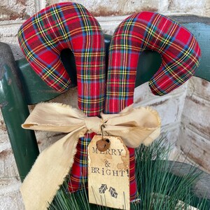 May include: Two fabric candy canes featuring a red, yellow, and blue plaid pattern. A beige frayed ribbon is tied around the center, holding a rectangular paper tag with the text MERRY & BRIGHT and two holly leaf icons, topped with a small metal bell.