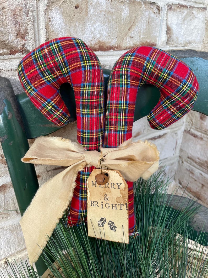 May include: Two fabric candy canes featuring a red, yellow, and blue plaid pattern. A beige frayed ribbon is tied around the center, holding a rectangular paper tag with the text MERRY & BRIGHT and two holly leaf icons, topped with a small metal bell.