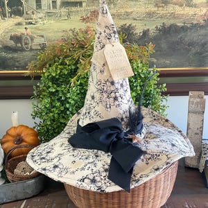 May include: A beige witch hat with a black floral pattern, a black bow, and a feather, resting on a woven basket. The hat has a tag that reads "Salem Hats Est. 1692". The hat is a decorative item.